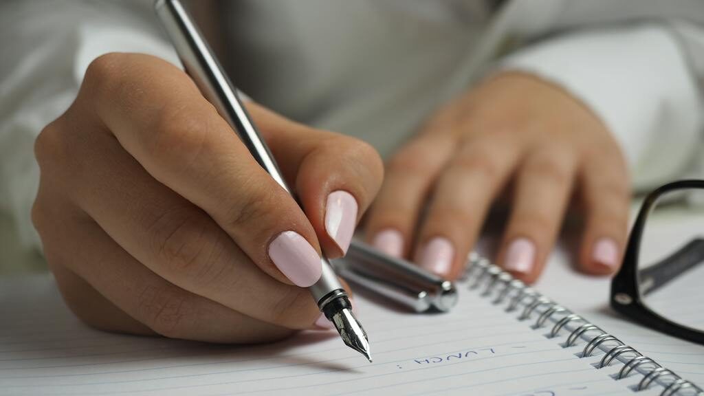 Woman in White Long Sleeved Shirt Holding a Pen Writing on a Paper